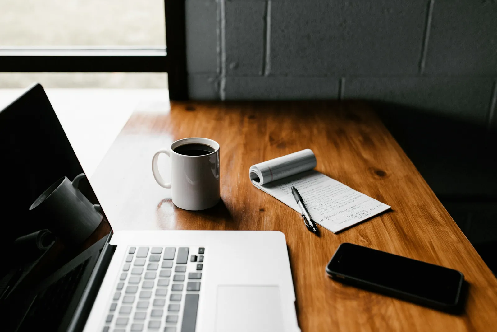 A wooden desk with a laptop, a notepad and a phone