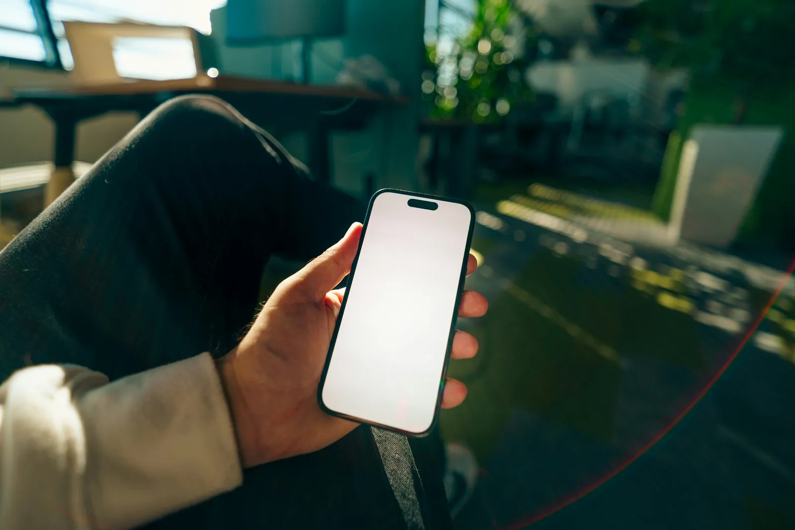 Une main tient un smartphone à l'écran blanc dans un espace de travail lumineux.