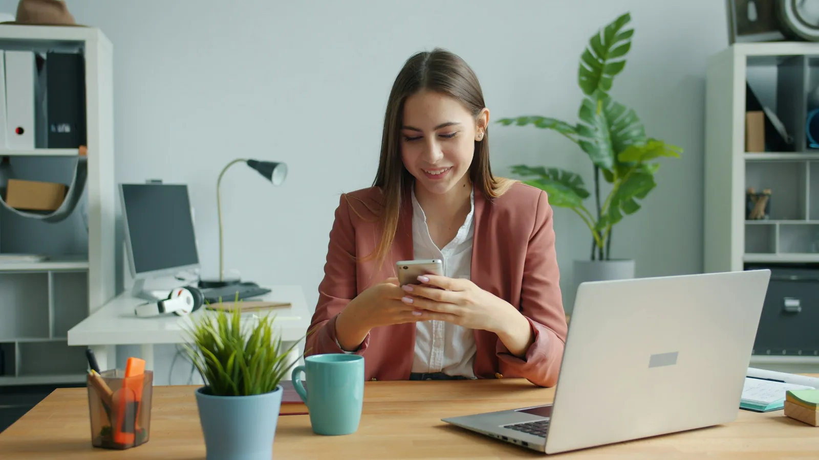 Une jeune femme en blazer consulte son smartphone, assise à un bureau devant un ordinateur portable.